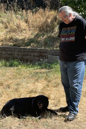 A tall white man stands in a back yard with dried-out grass. He looking at a black dog lying on the ground next to him, and positioning himself to shade the dog with his shadow. The man has a long grey ponytail and glasses, and is wearing a jeans, a black t-shirt, and black sneakers. The t-shirt has all-caps text multiple lines of different colors with the statements 'Black Lives Matter', 'We don't need a wall', 'News isn't fake', 'Science is real', 'No human is illegal', 'Women's rights are human rights', 'Love is love', and 'Kindness is everything'.