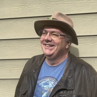 A white man with large glasses and a tan-grey, wide-brimmed fedora smiles and looks up and to the left. He is wearing a blue t-shirt and a dark brown leather working jacket. There are pins attached to his hat. 